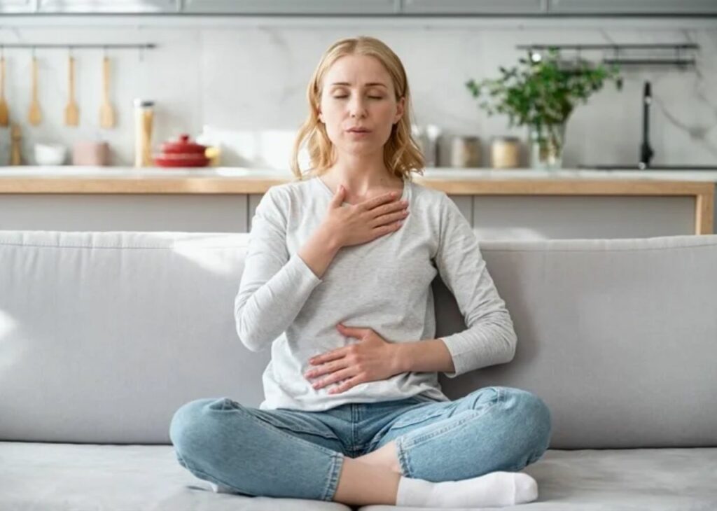 Woman practicing deep breathing exercises with one hand on her chest and the other on her abdomen, sitting on a couch in a calm home setting.