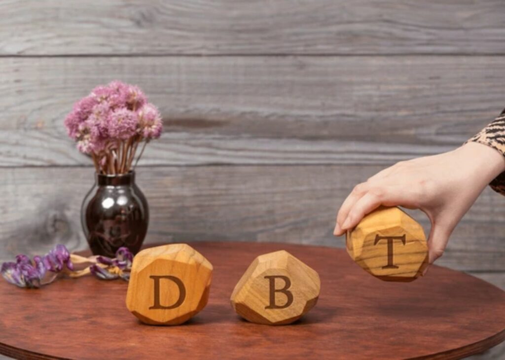 The image shows three wooden blocks, each with a letter engraved on them, spelling out "DBT." A hand is shown adjusting or placing the "T" block, while the other two blocks with "D" and "B" rest on a wooden surface. In the background, there is a dark-colored vase with light pink flowers and a rustic wooden backdrop. This arrangement suggests the concept of Dialectical Behavior Therapy (DBT), a therapeutic approach often used for emotional regulation and treating mental health disorders like borderline personality disorder. The calm setting may also emphasize the soothing and therapeutic nature of DBT.