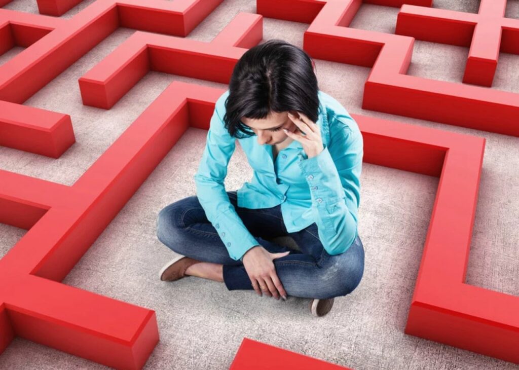 Frustrated woman sitting in a maze with red walls, holding her head in her hand, symbolizing confusion or feeling stuck