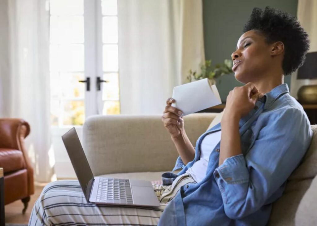 Woman sitting on a couch with a laptop on her lap, holding an envelope and fanning herself, appearing warm in a bright living room.