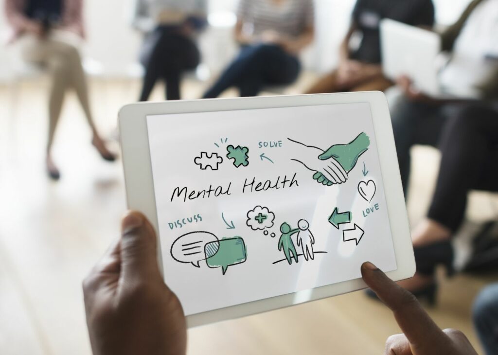 Person holding a tablet displaying a mental health diagram with words like 'discuss,' 'solve,' and 'love,' during a group training session.