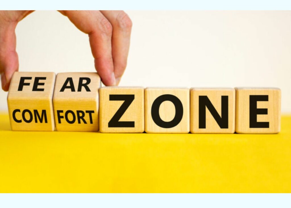 The image shows wooden blocks arranged to spell out the words "FEAR ZONE" and "COMFORT ZONE." The blocks are positioned so that one block can be rotated to change between the words "FEAR" and "COMFORT," implying a transition between fear and comfort. This visual represents the idea of moving from a "comfort zone" to a "fear zone" or vice versa, symbolizing personal growth or stepping out of one's comfort zone to face challenges and new experiences.